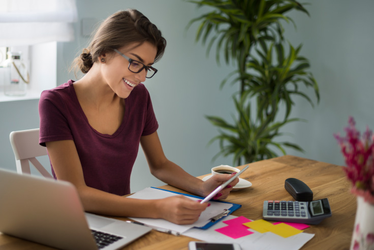 Cayman Branch la mejor opción para administrar tu dinero Mujer contenta con tablet en mano revisando informe, cuentas; laptop, calculadora, post it y taza de café sobre escritorio