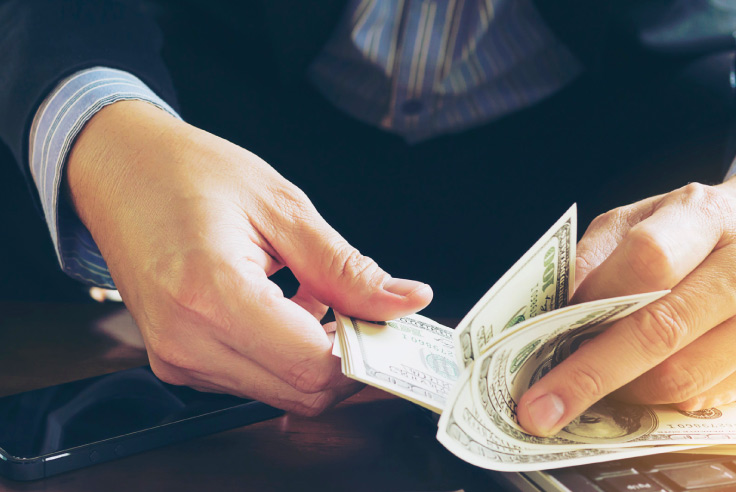 Hombre con dinero en mano contando los billetes en dólares sobre laptop y télefono