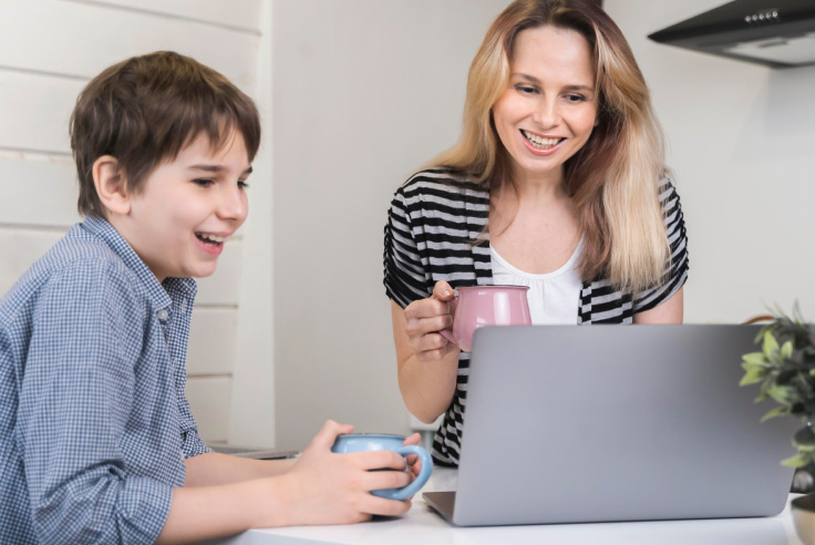 Niño con taza azul en la mano y mamá con taza rosada, ambos sonrientes, viendo la pantalla de laptop