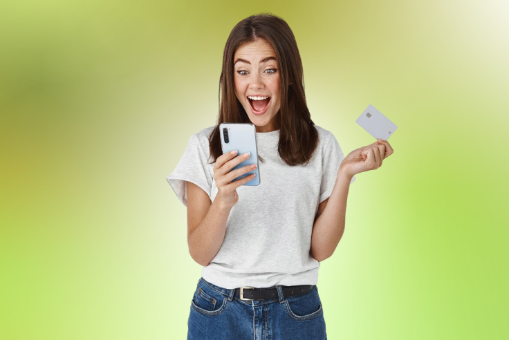Mujer joven sorprendida mirando su teléfono con una Tarjeta con tecnología contactless en la mano