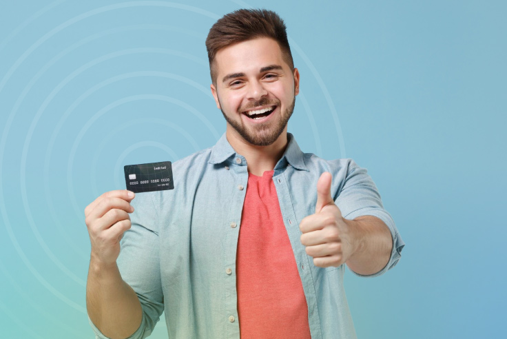 Joven sonriente con barba sostiene tarjeta bancaria en la mano y hace señal de aprobación con la otra promoviendo seguridad